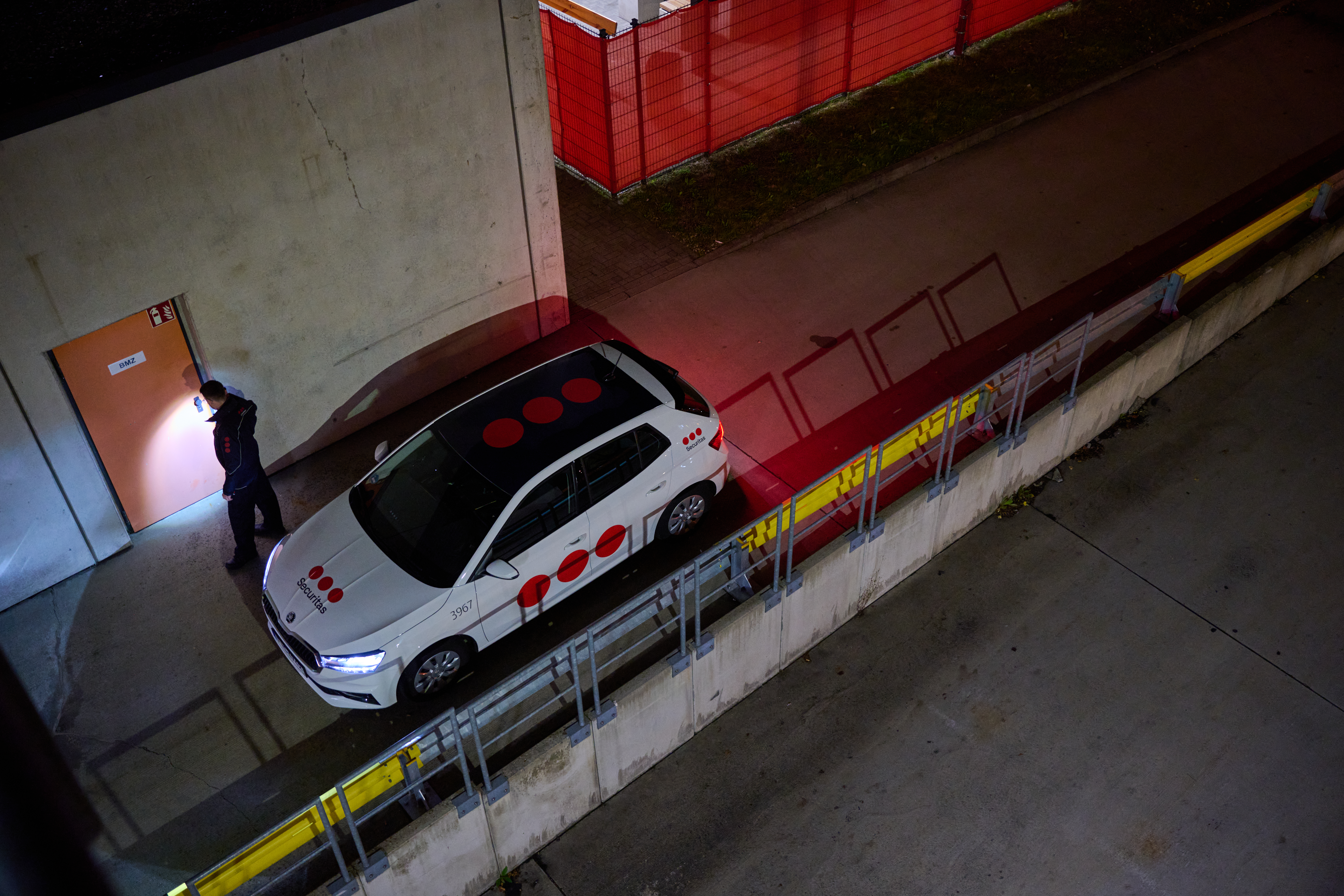 Overhead view of a Securitas guard securing the outdoors of a facility with a Securitas car parked next to him.